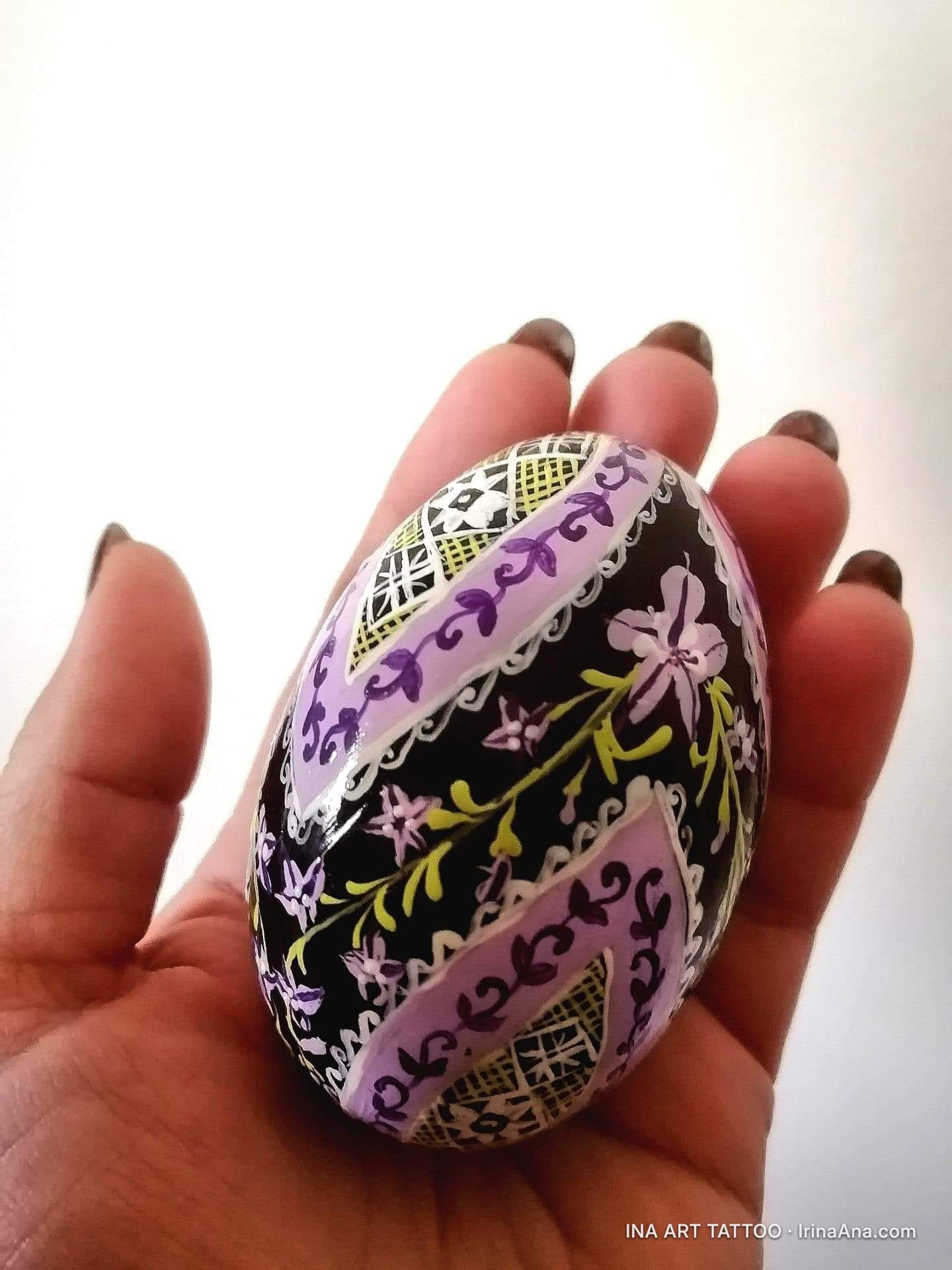 O mână ține un ou încondeiat cu modele florale și geometrice în nuanțe de violet, negru, alb și galben.