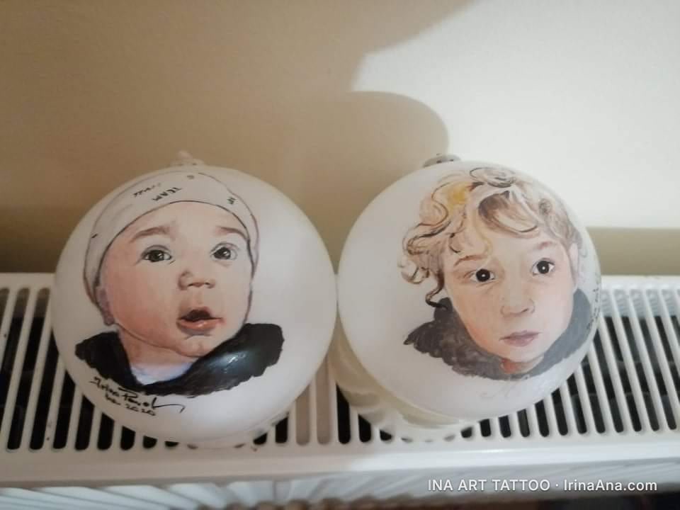 Două ornamente albe de Crăciun cu portrete pictate de copii, așezate pe un calorifer alb.