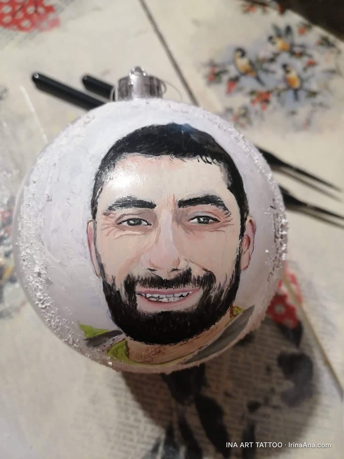 Un portret pictat al unui bărbat zâmbitor, cu barbă, pe o decorațiune albă de Crăciun.