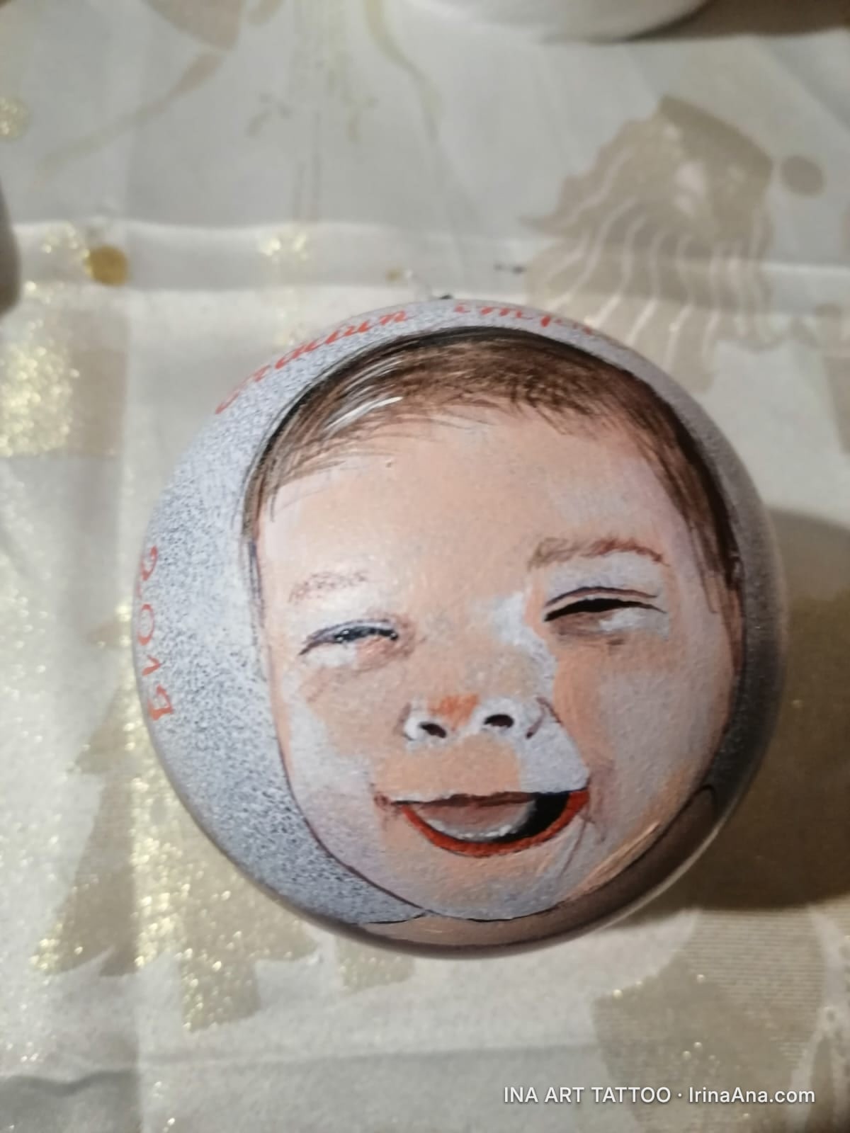 Un glob de Crăciun pictat manual, cu un portret de bebeluș zâmbitor, pe un fundal alb-sclipitor.