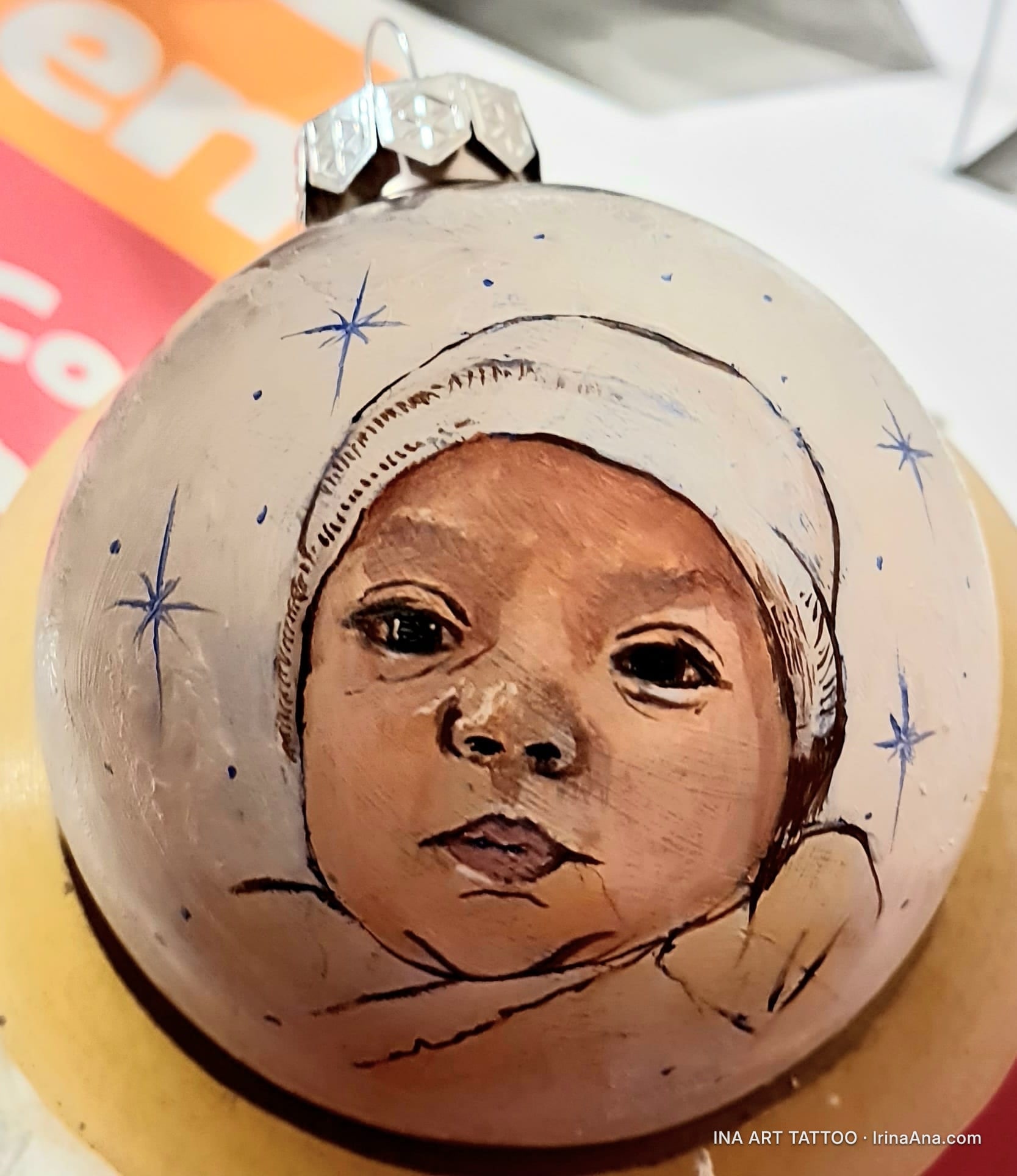 Un glob de Crăciun alb, pictat manual, cu un portret de bebeluș cu căciuliță albă, înconjurat de steluțe albastre.
