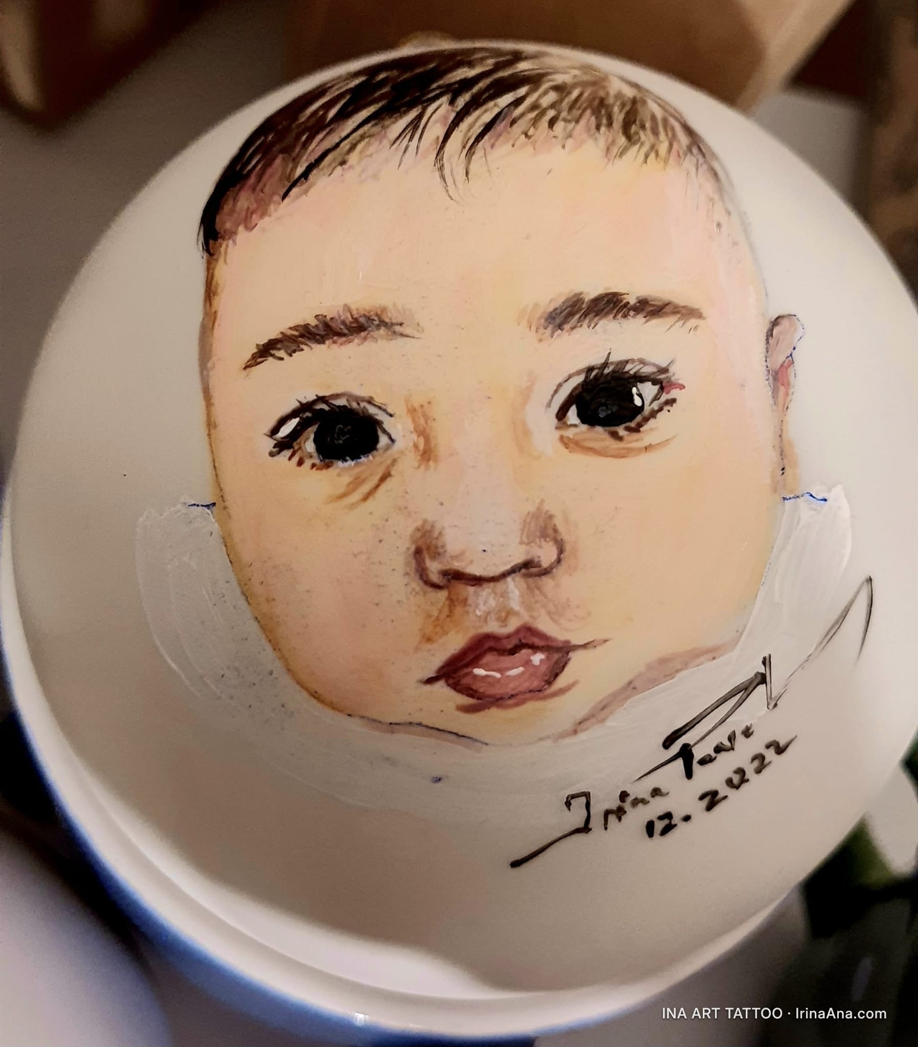 O decorațiune rotundă de Crăciun, pictată manual cu un portret de bebeluș cu ochi mari, negri și păr închis la culoare.