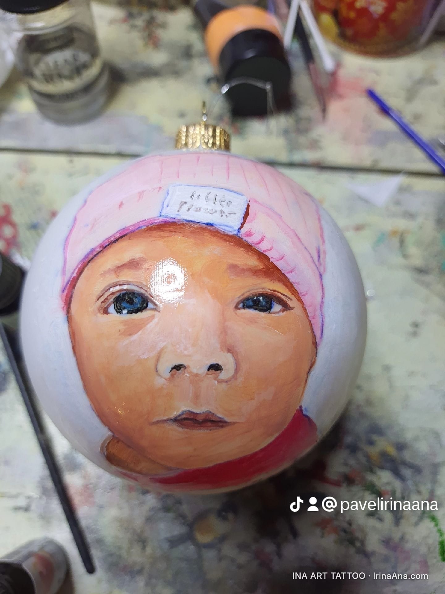 Un ornament de Crăciun pictat manual, cu un portret detaliat al unui bebeluș cu o căciulă roz și ochi albaștri.