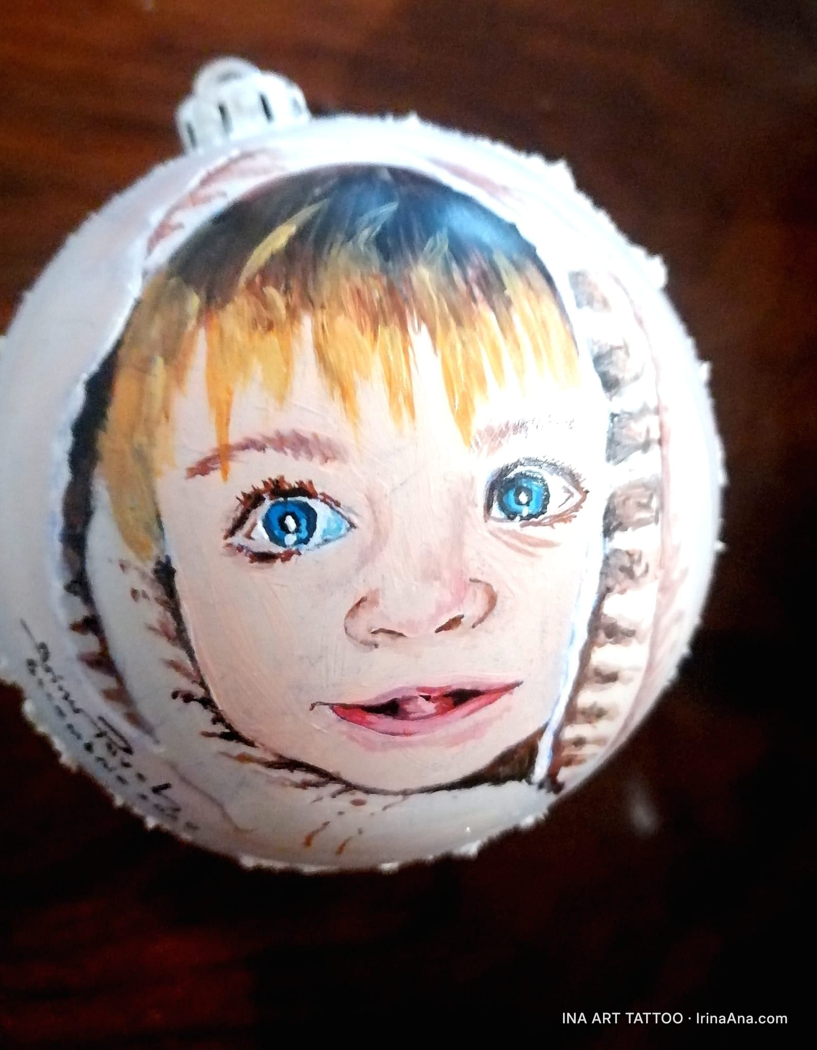 Un glob de Crăciun alb, pictat manual cu un portret de copil cu păr blond și ochi albaștri, pe un fundal maro închis.
