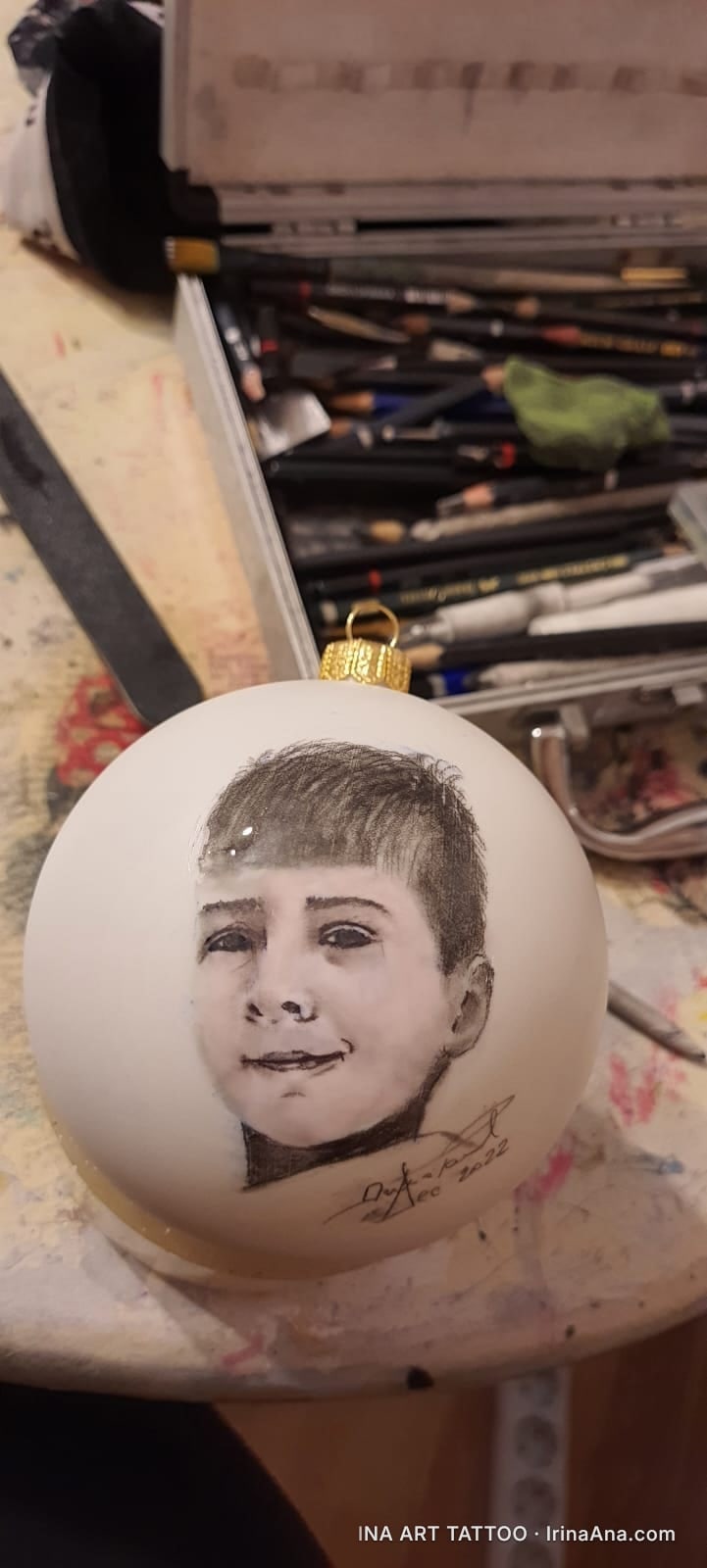 Un glob de Crăciun alb, mat, cu un portret desenat în creion al unui copil zâmbitor. În fundal, o cutie cu creioane.