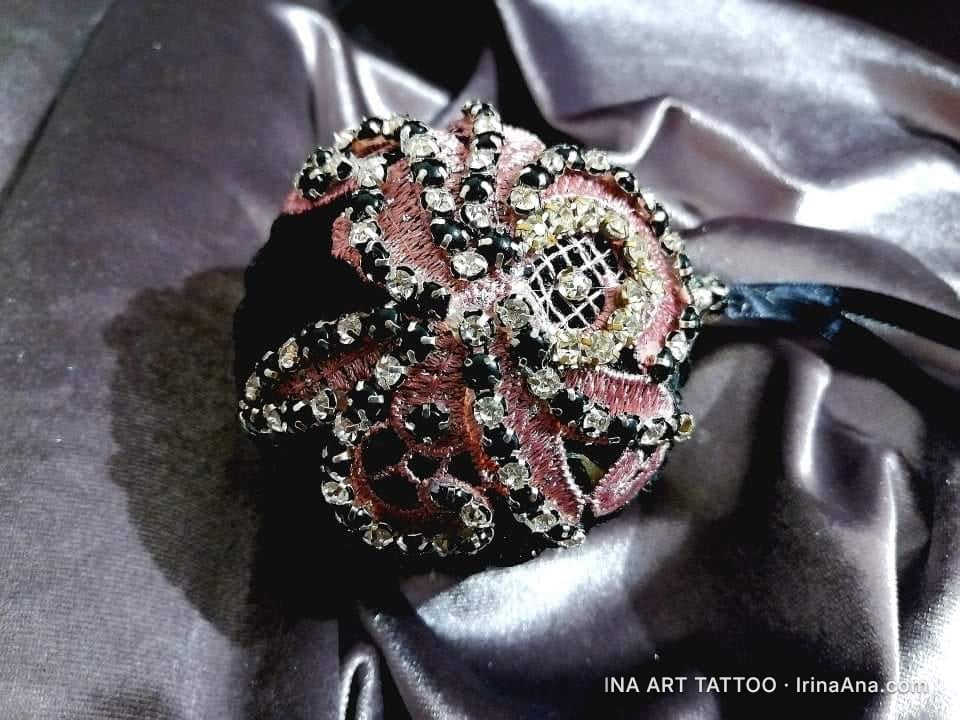 O floare decorativă brodată, cu petale rozalii și negre, împodobită cu strasuri strălucitoare, așezată pe o draperie de sati…