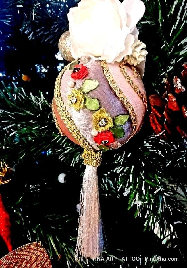 Un ornament de Crăciun lucrat manual, sferic, decorat cu panglici, flori textile și un ciucure, atârnat într-un brad.
