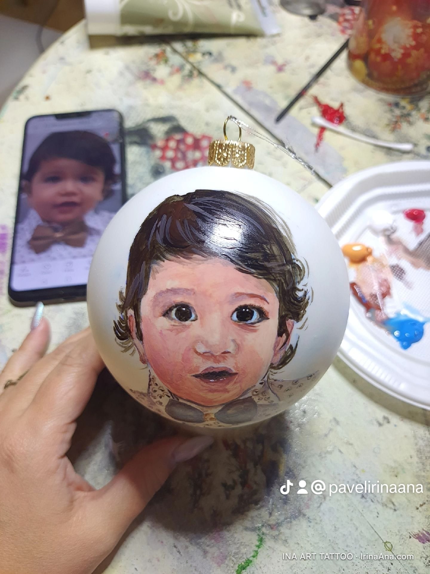 O mână ține un glob de Crăciun alb cu un portret pictat al unui copil, lângă un telefon cu fotografia sursă.