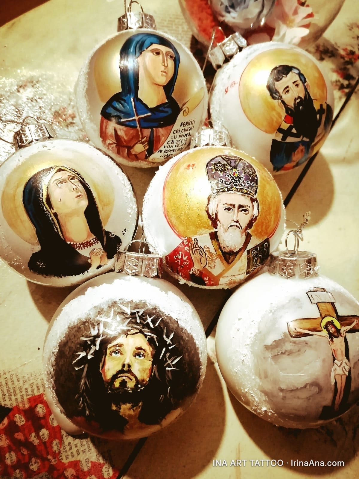 Șase globuri de Crăciun albe, pictate manual cu scene și figuri religioase ortodoxe, așezate pe o suprafață deschisă.