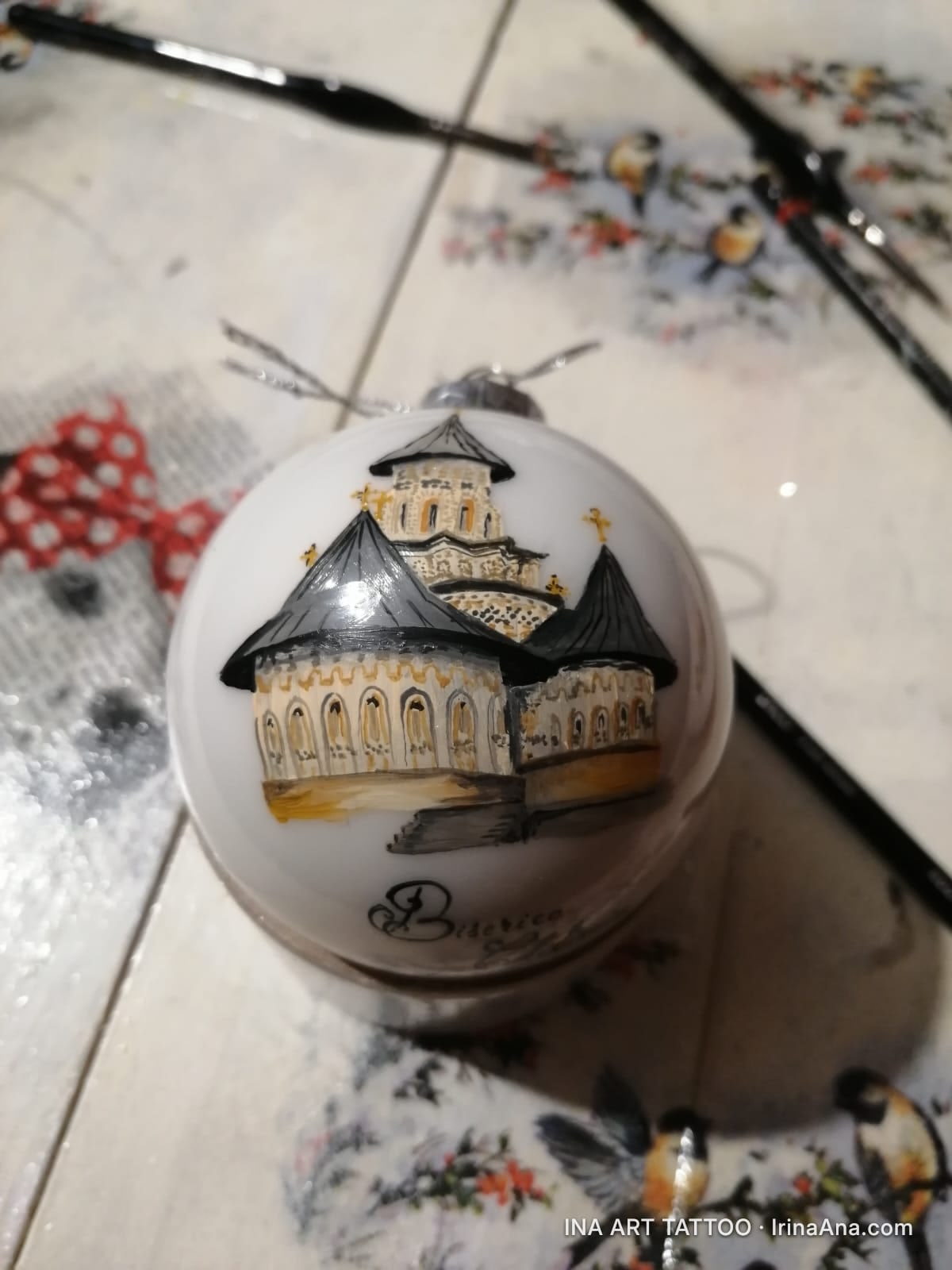 Un glob de Crăciun alb, pictat manual cu o mănăstire ortodoxă cu acoperișuri negre și cruci aurii, pe o masă.