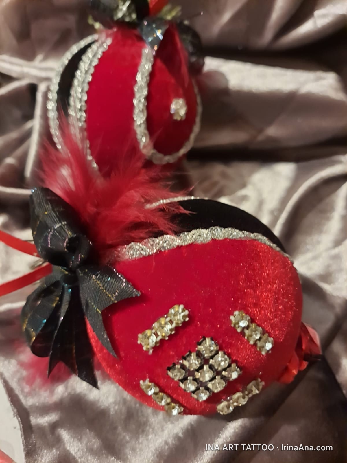 Două ornamente de Crăciun din catifea roșie, decorate cu pene roșii, panglici negre și ștrasuri argintii, pe un fundal gri.