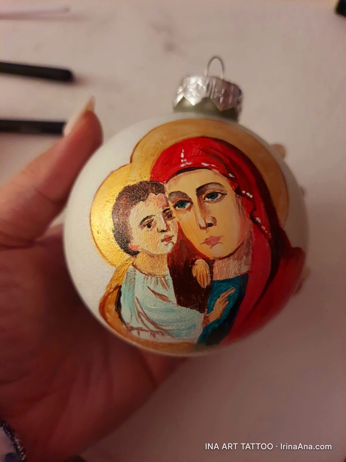 Un glob de Crăciun alb, pictat cu o icoană religioasă: Fecioara Maria cu pruncul Iisus. Globul este ținut într-o mână.