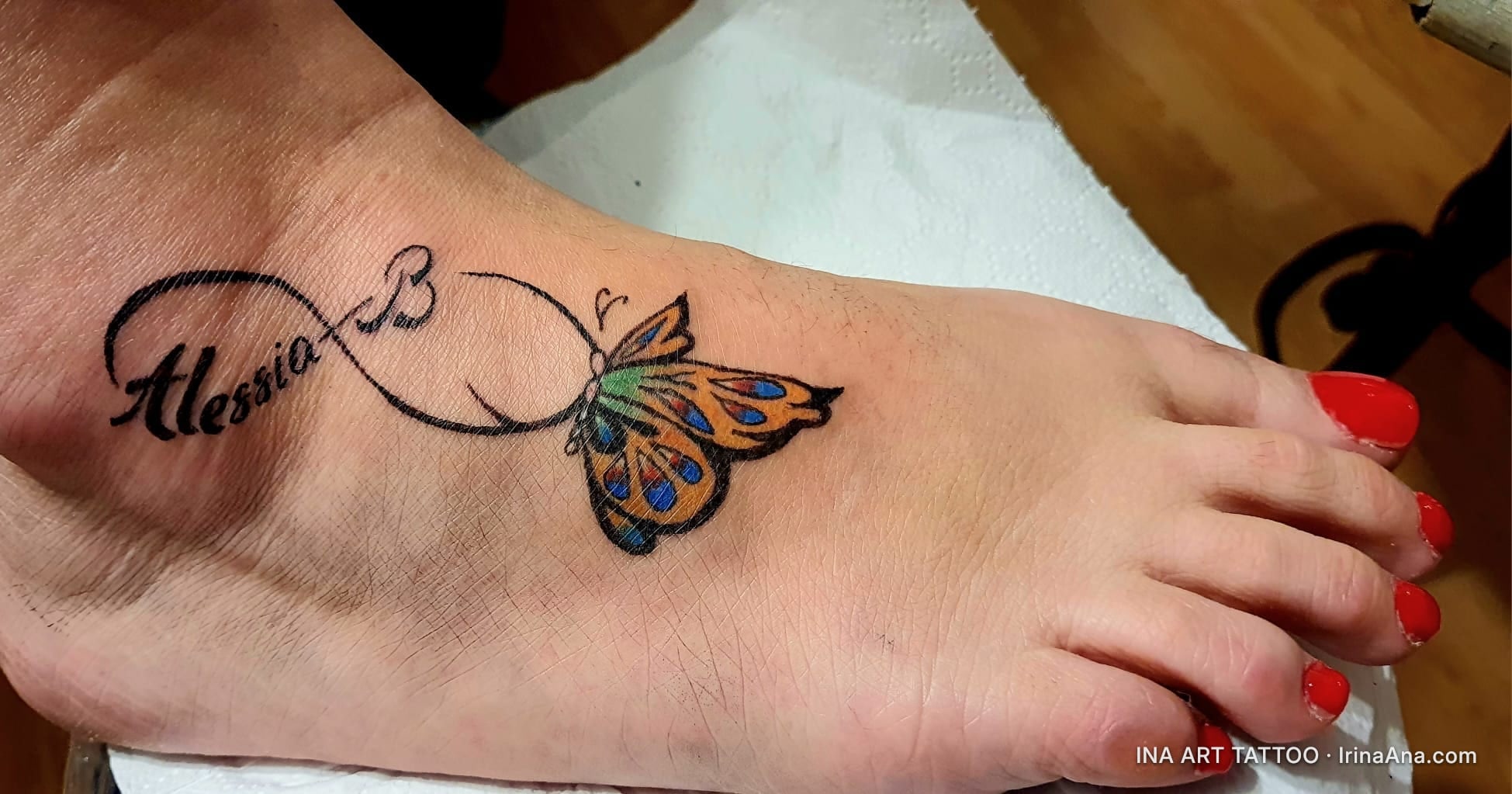 Colored tattoo on foot, featuring an infinity symbol, the name Alessia, the letter B, and a butterfly with orange and blue w…