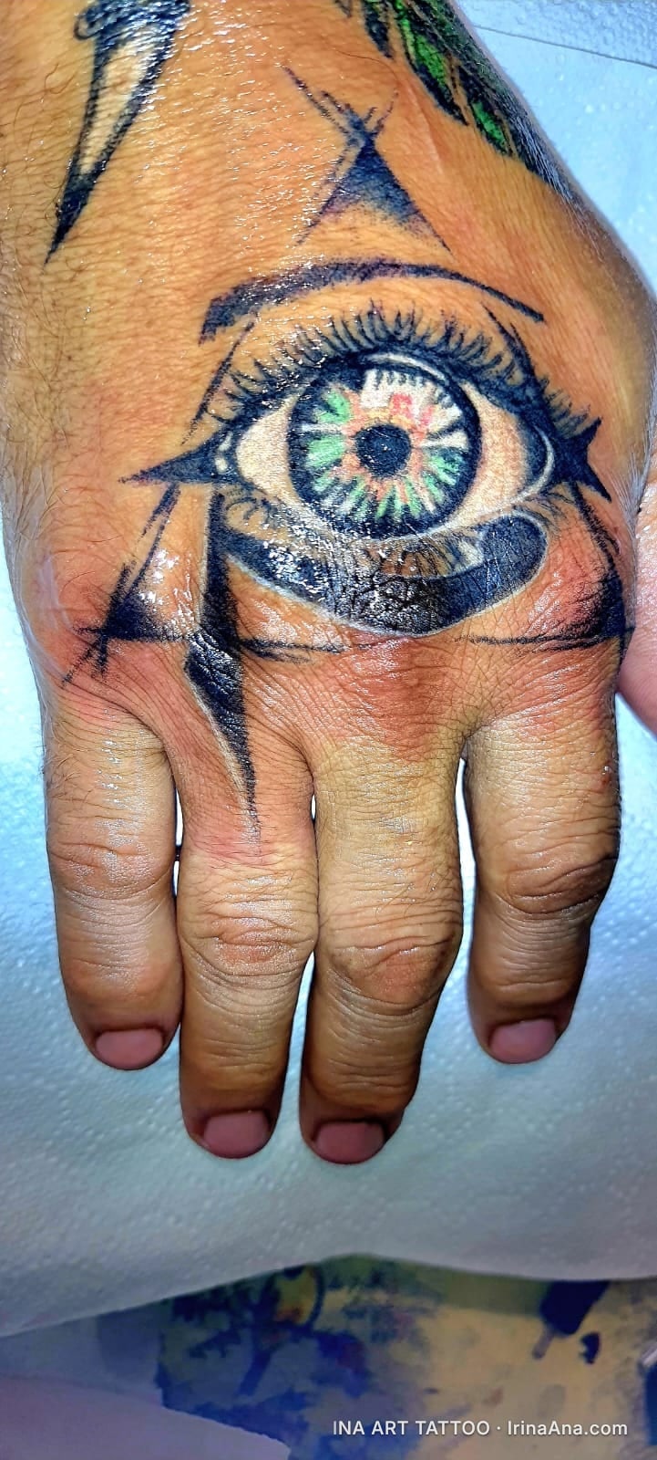 Colorful all-seeing eye tattoo with green and black iris, on the back of the right hand.