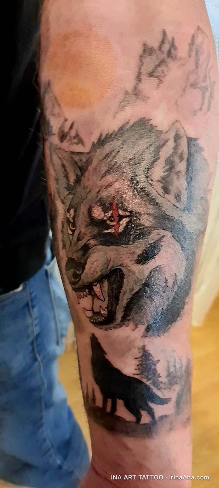 Forearm tattoo of an angry gray wolf with an open mouth and a red eye scar, above a howling black wolf, against a forest and…