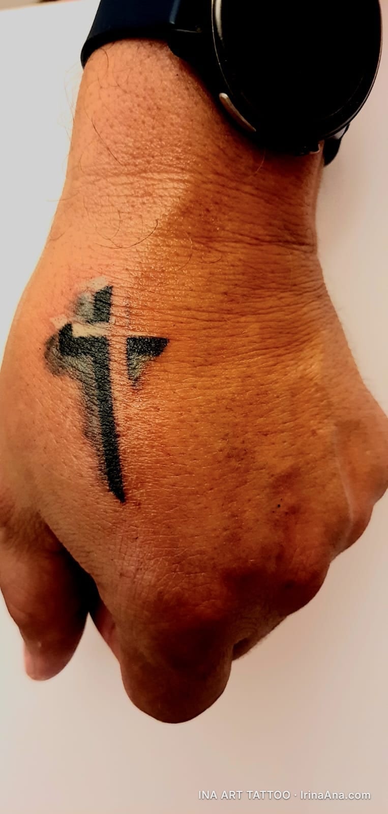 Tattoo of a stylized black cross on the back of a person's right hand, with a black watch on the wrist.