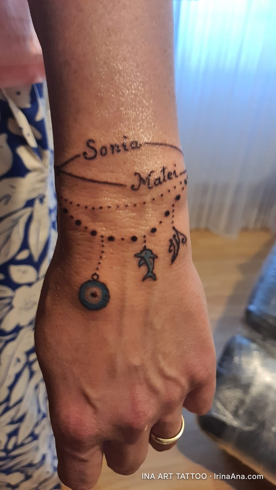 Wrist tattoo featuring the names Sonia and Matei, a string of beads, and three charms: a blue evil eye, a dolphin, and a tri…