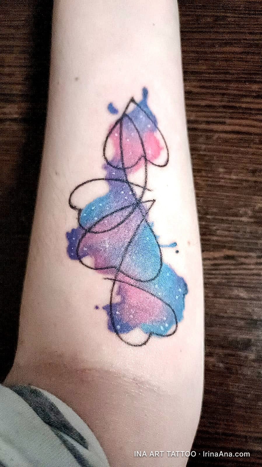Forearm tattoo of abstract, galaxy-colored hearts in blue, pink, and purple, on light skin.