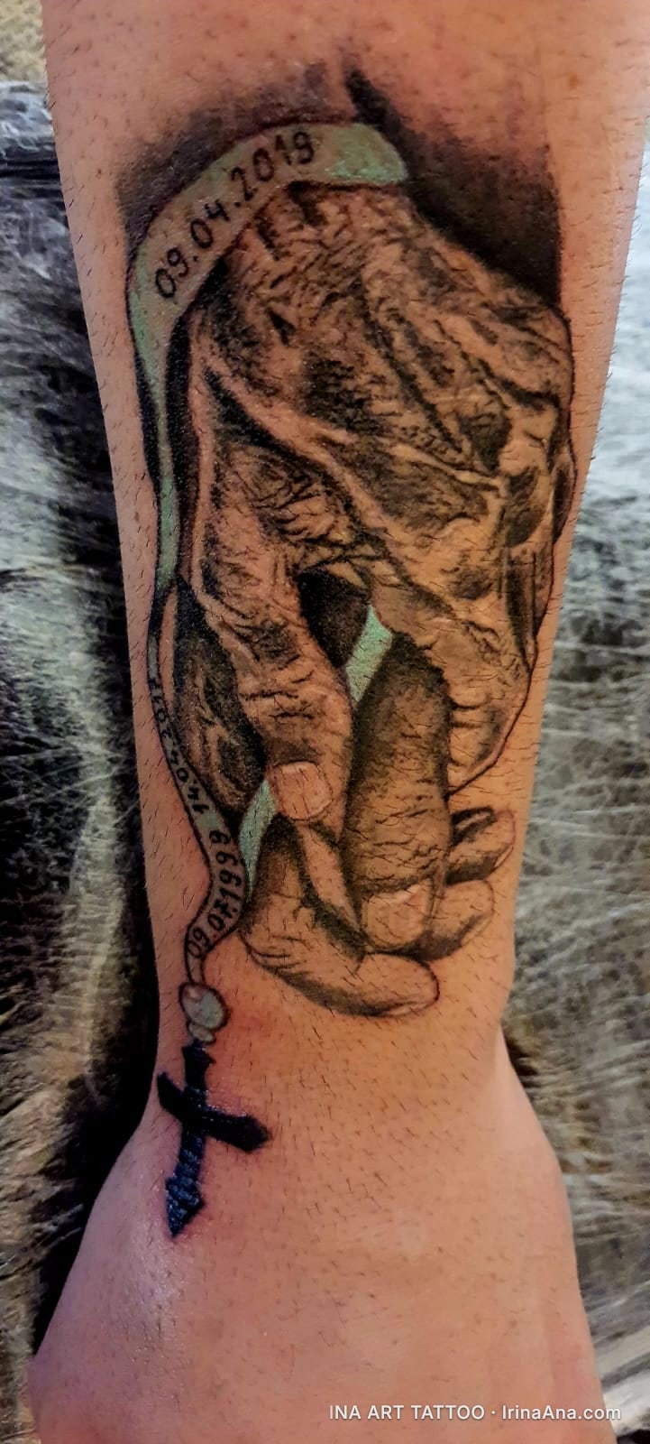 Realistic tattoo on forearm depicting two hands, one adult and one child, holding each other. A rosary with a cross and date…