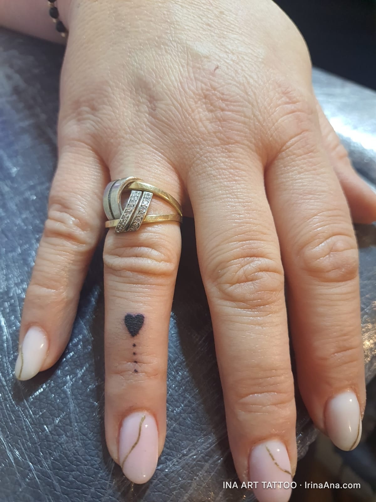 Minimalist tattoo of a black heart and three dots below it, on the ring finger of a hand with nude manicure.