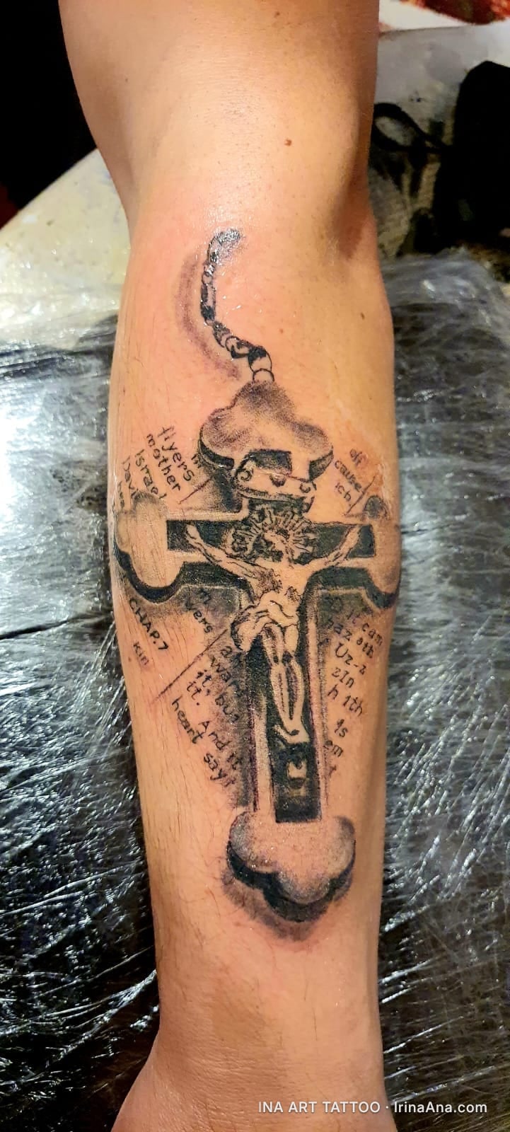 Black and white tattoo on forearm, depicting a crucifix with Jesus, a crown above, and fragments of religious text.