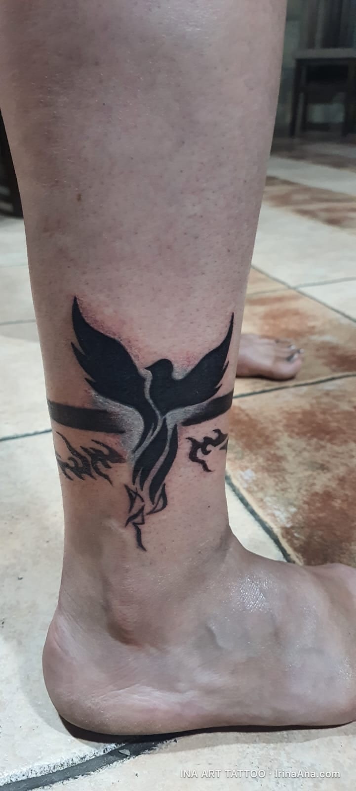 Monochrome ankle tattoo depicting a phoenix bird with outstretched wings, surrounded by stylized flames.