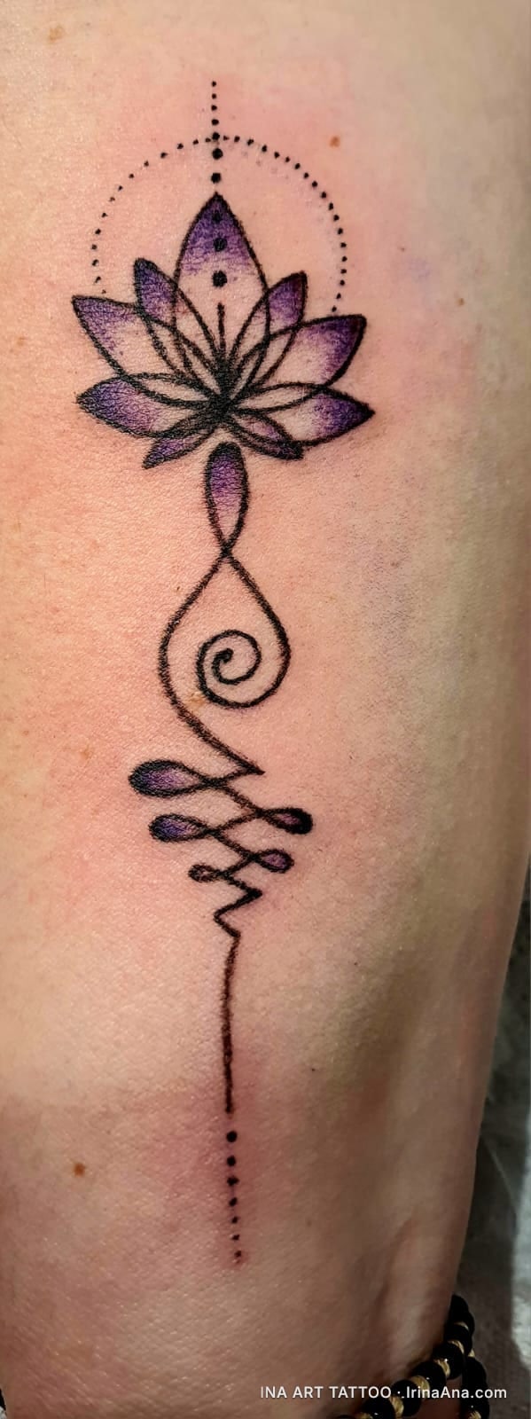 Vertical tattoo on skin of a purple lotus flower with an unalome symbol beneath it, surrounded by black dots.