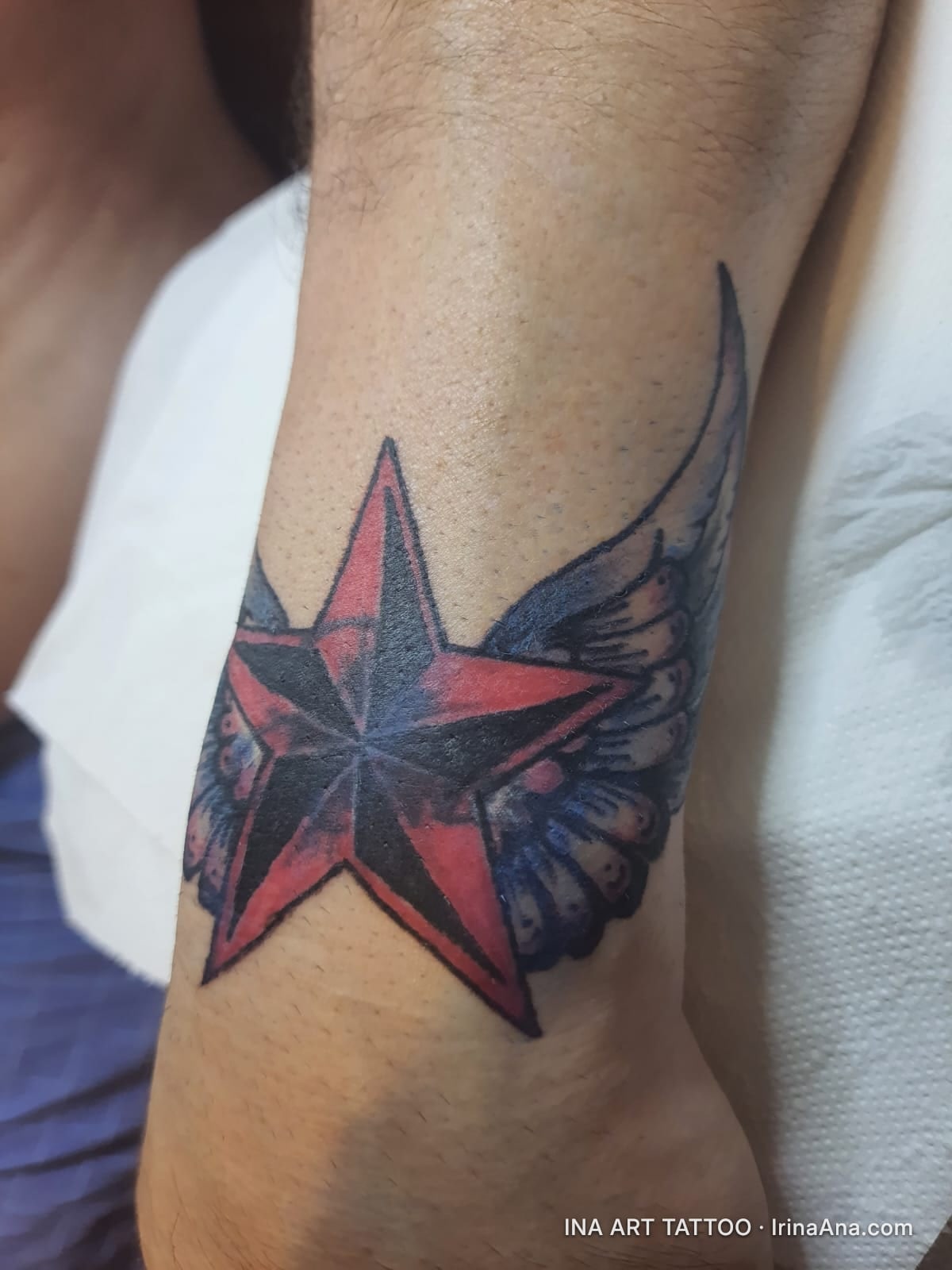 Colored tattoo of a red and black star with blue wings on the wrist.