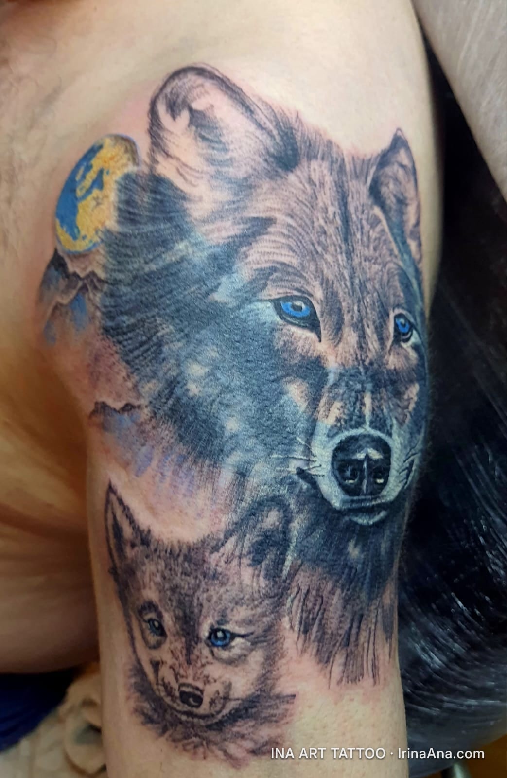 Color tattoo on shoulder with an adult wolf and a wolf cub, both with blue eyes, against a mountain background with a yellow…