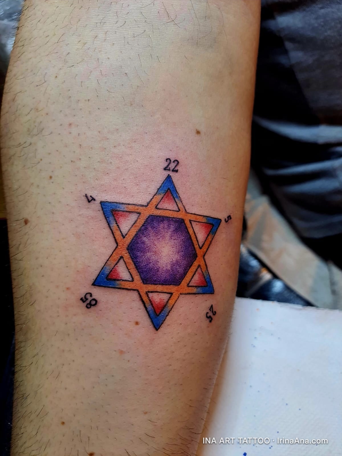 Colorful six-pointed star tattoo with an orange outline and blue and red shading, on a person's forearm. In the center, a pu…