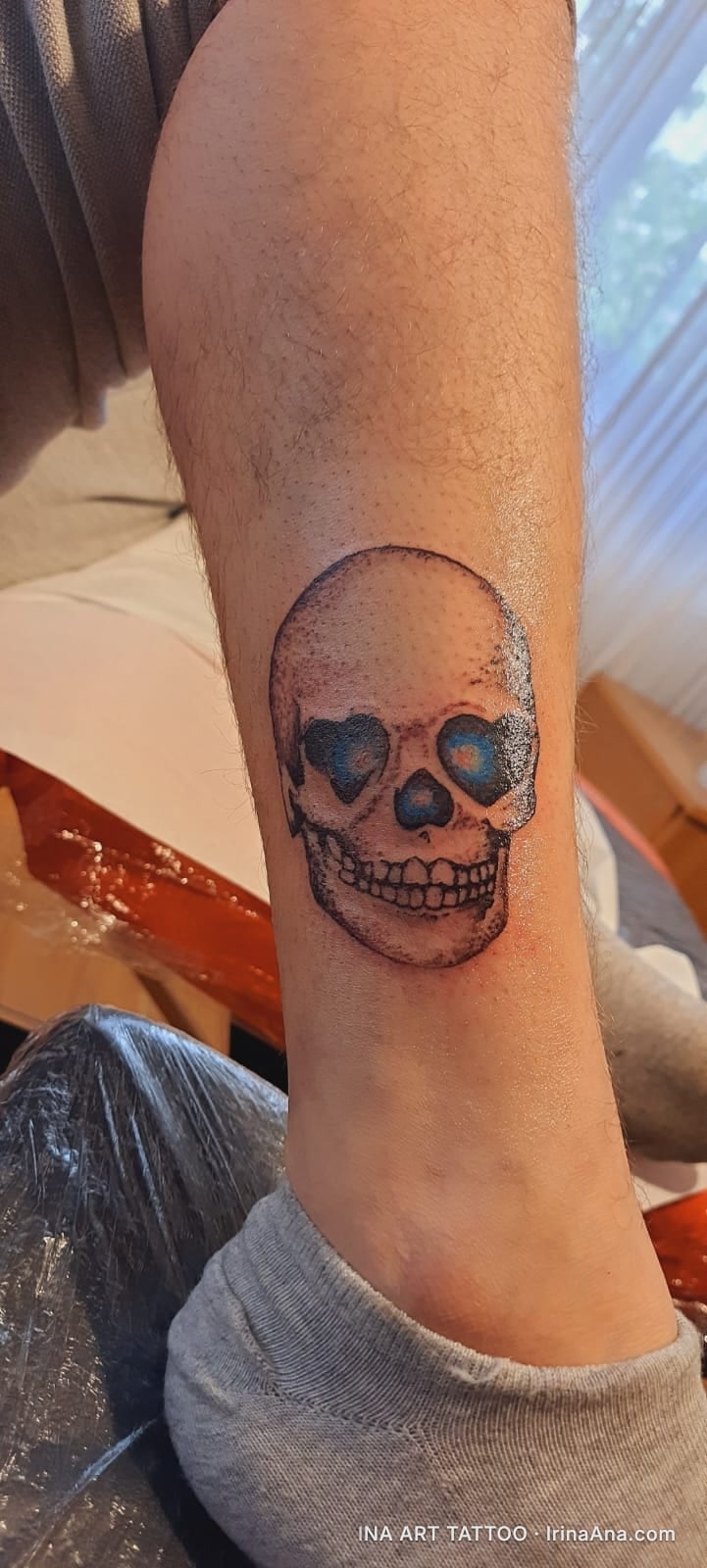 Tattoo on a calf of a skull with blue heart-shaped eye sockets, a black nose, and white teeth.