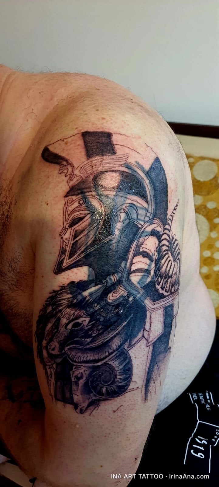 Black and white tattoo on a man's shoulder, depicting a Spartan helmet with ram horns and armor.