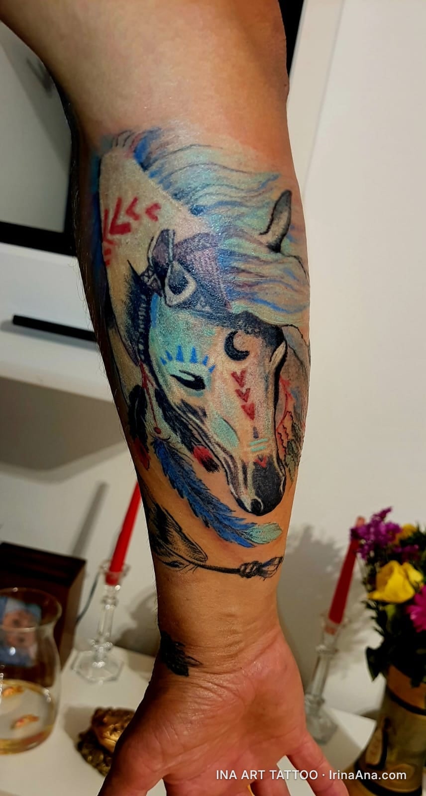 Colorful stylized horse head tattoo with blue and red feathers on a person's forearm.
