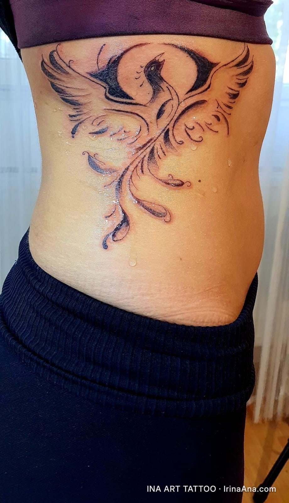 Tattoo of a stylized phoenix with wide-spread wings and decorative tail, on the side of the torso.