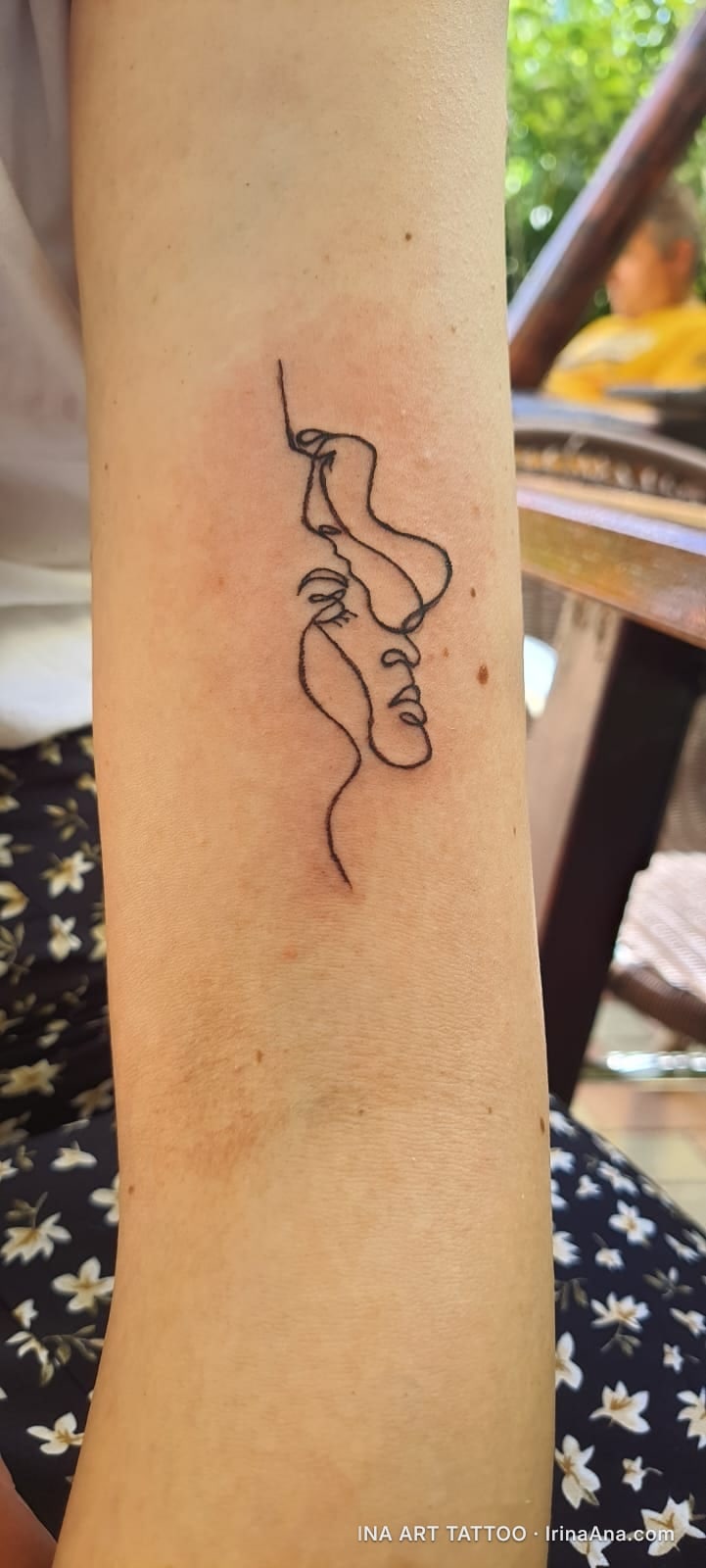 Forearm tattoo of two abstract faces, drawn with a single continuous line, their noses almost touching.