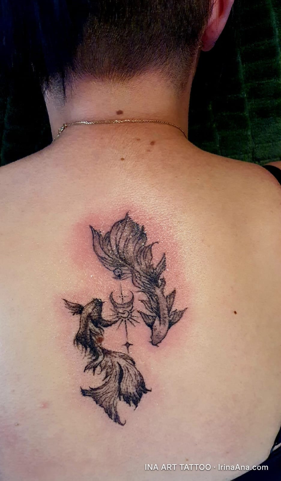 Black and white tattoo of two fish, a crescent moon, and stars, on a person's back.