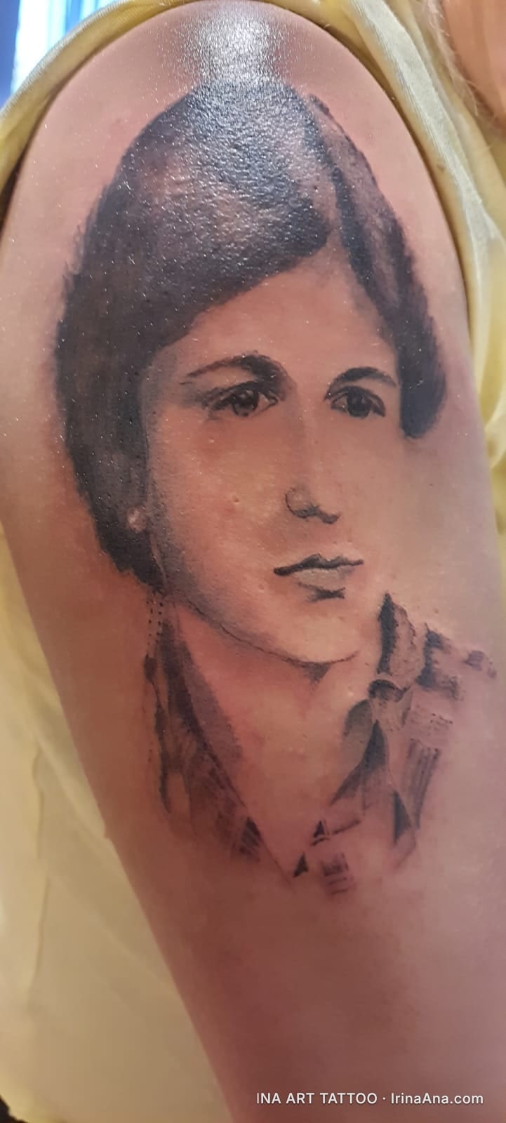 Black and white tattoo on arm, portrait of a young woman with dark hair and a plaid shirt.