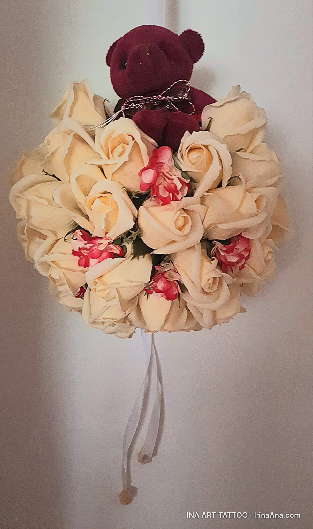 A round bouquet of cream roses and red flowers, with a dark brown teddy bear in the center and white ribbons hanging.