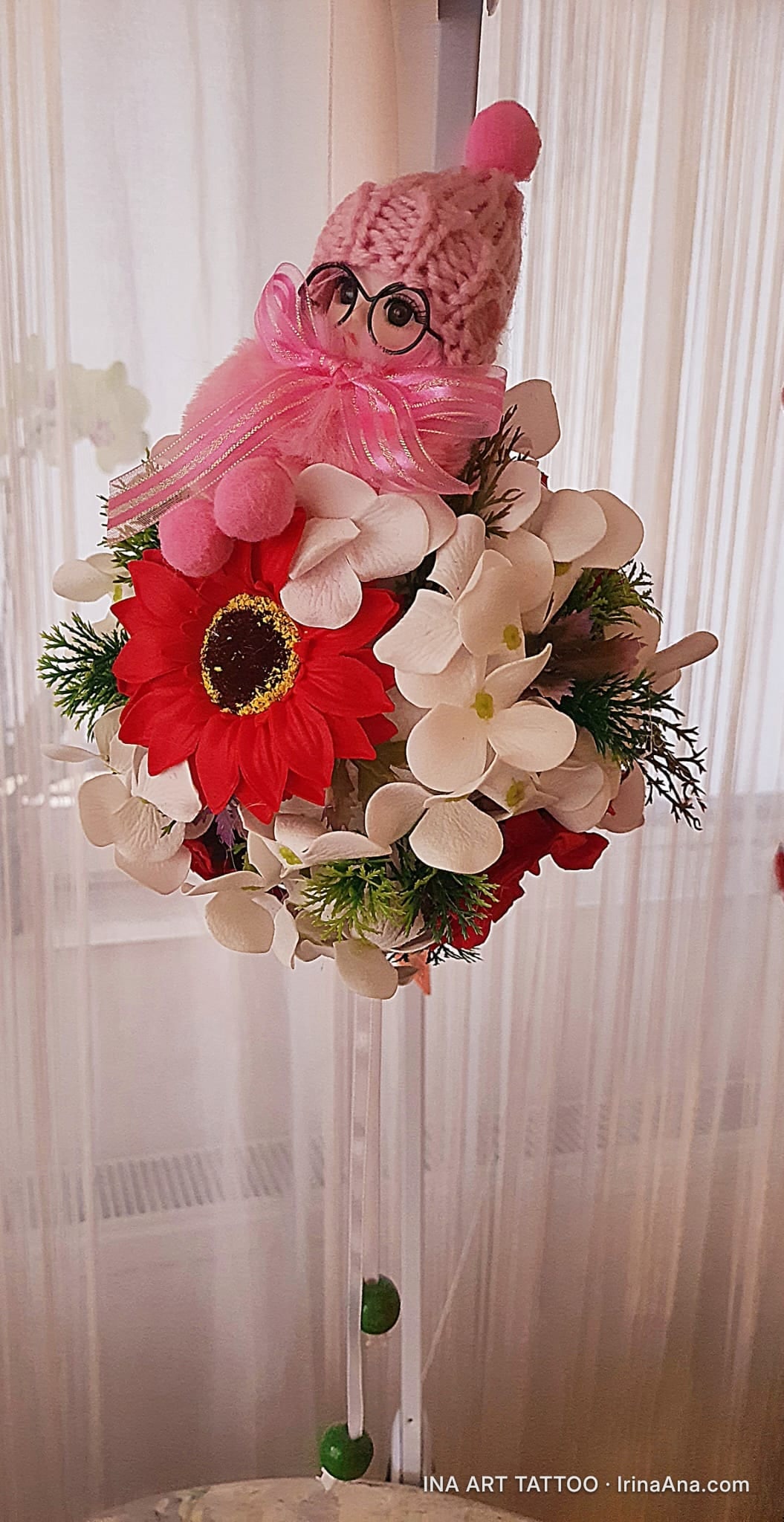 Floral arrangement with a pink doll wearing glasses and a knitted hat, placed on a tall stand, in front of a white curtain.