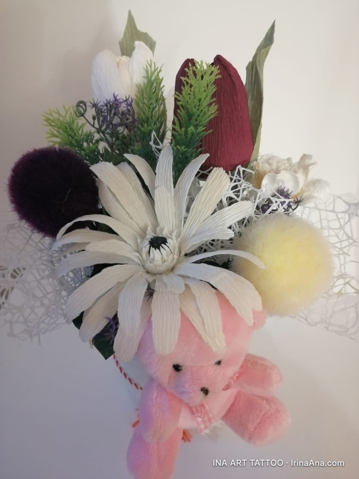 Crepe paper flower arrangement with white, red, and green flowers, and a pink plush teddy bear hanging.