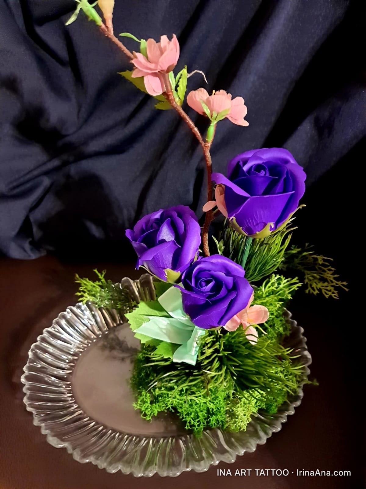 Handmade floral arrangement with three purple roses, pale pink blossoms, and green moss, in an oval glass tray.