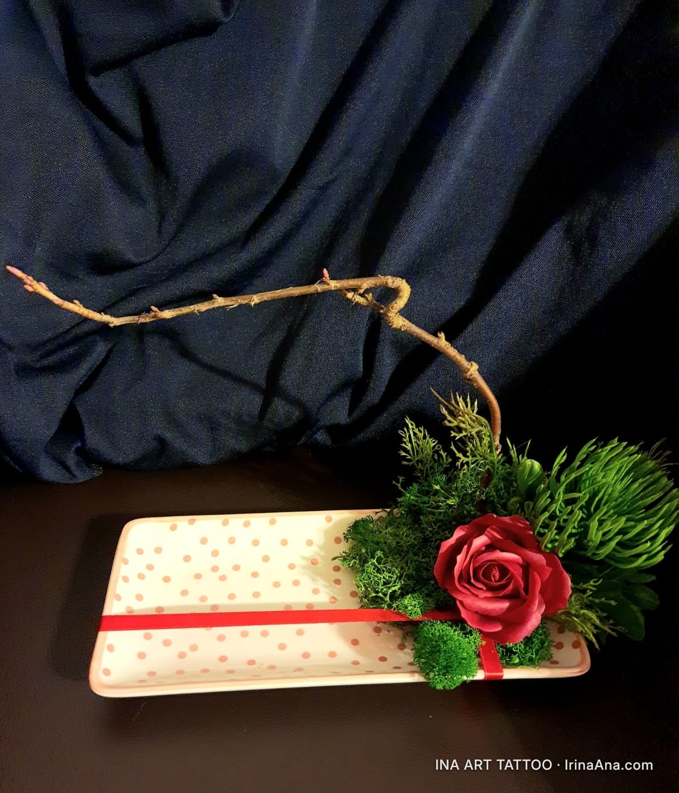 Crepe paper floral arrangement with a red rose, green moss, and a branch, on a white tray with pink polka dots.