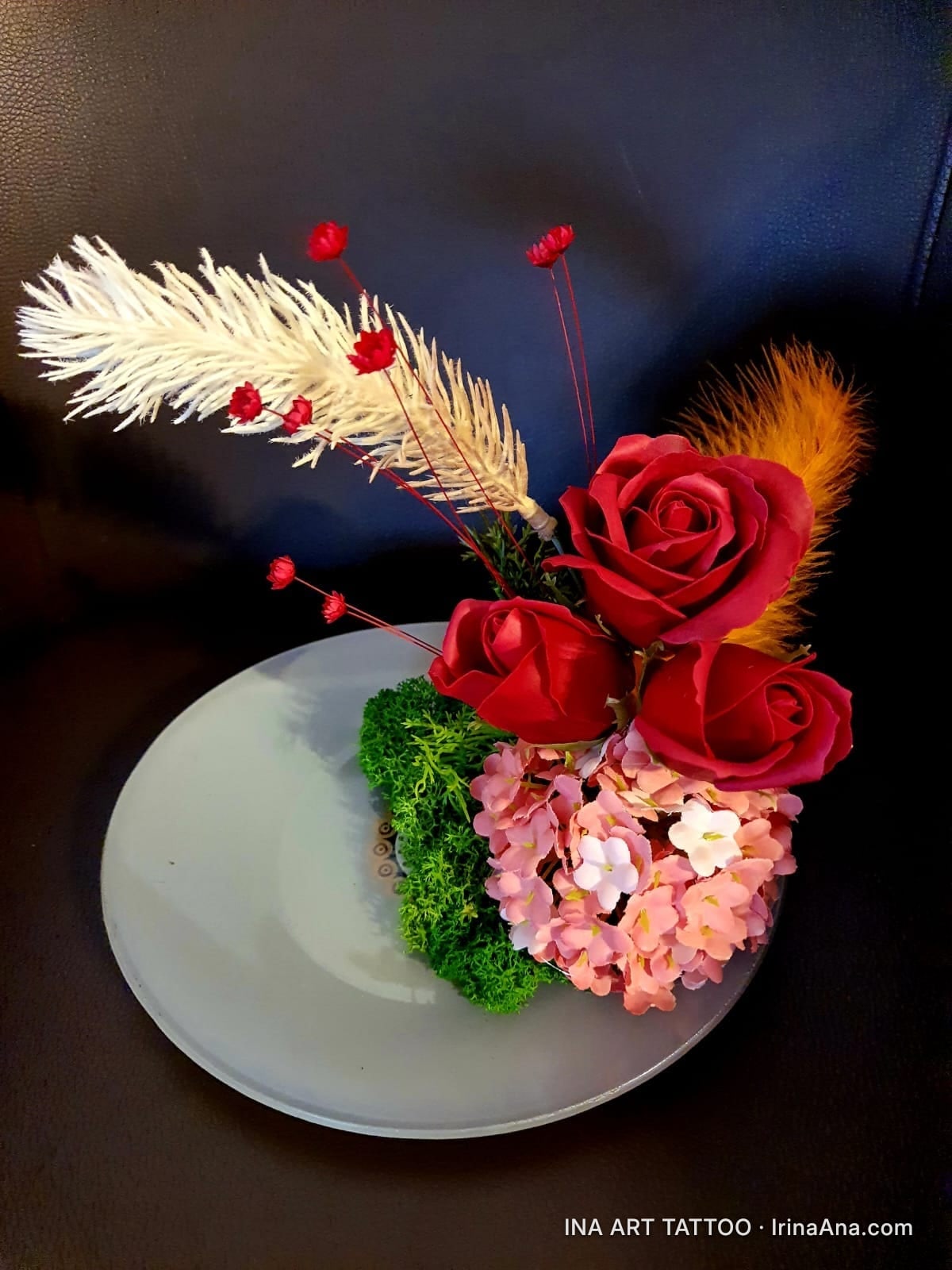 Handmade floral arrangement with three red roses, pink flowers, green moss, and white feathers, on a gray plate.
