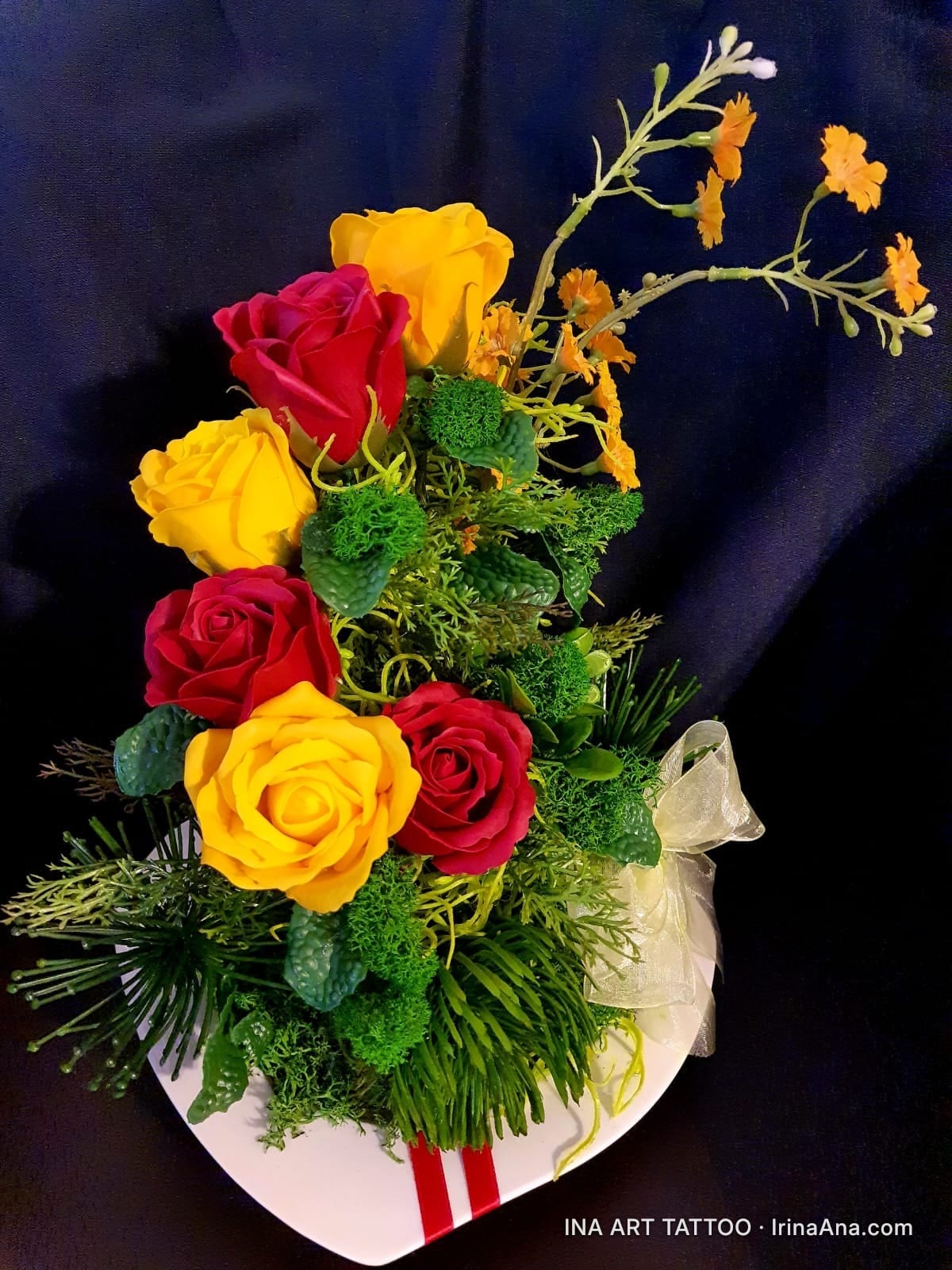 Floral arrangement with red and yellow roses, green plants, and small orange flowers, in a white vase.