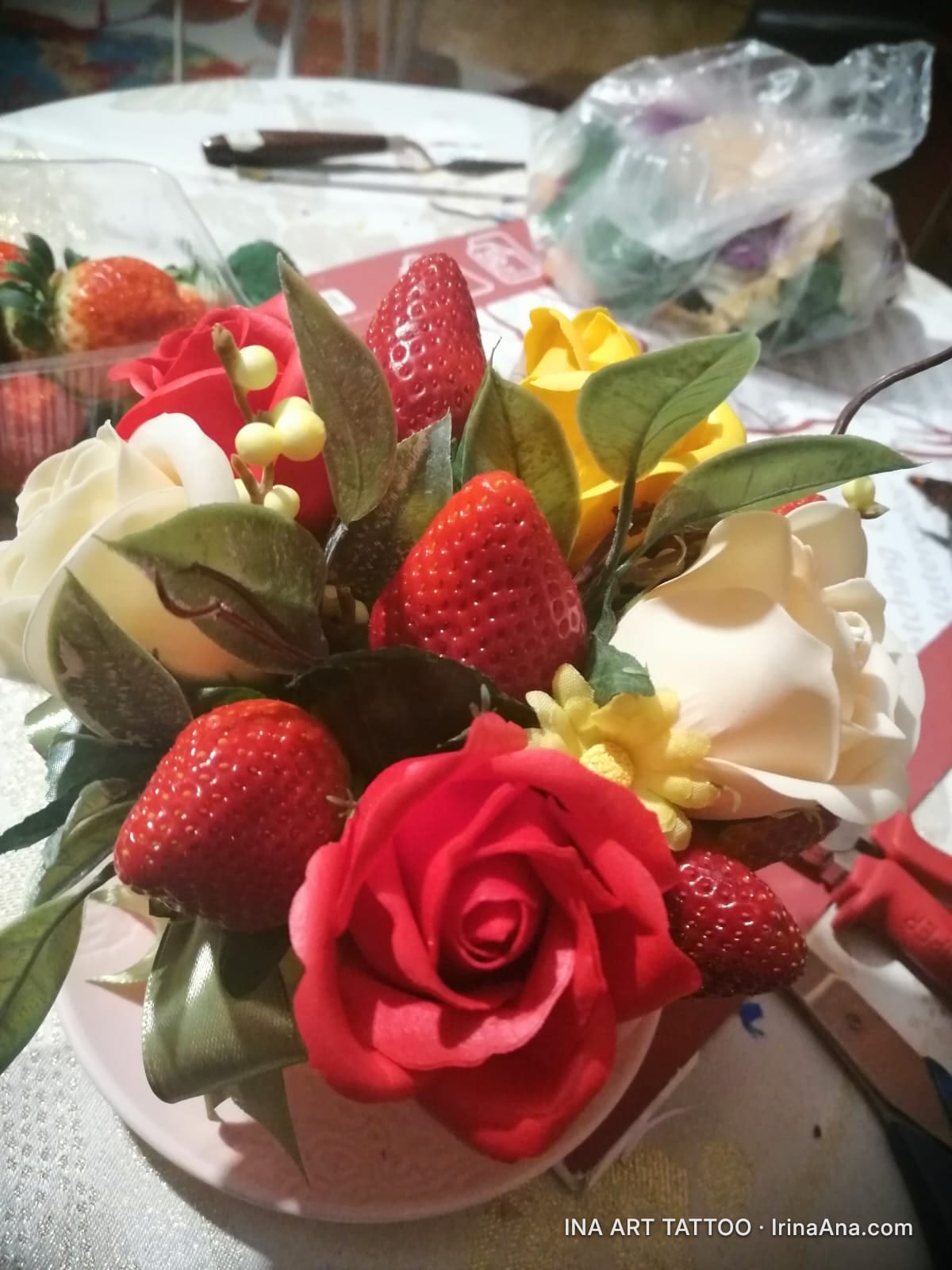 Hand-made floral arrangement with red, yellow, and white crepe paper roses, green leaves, and strawberries.