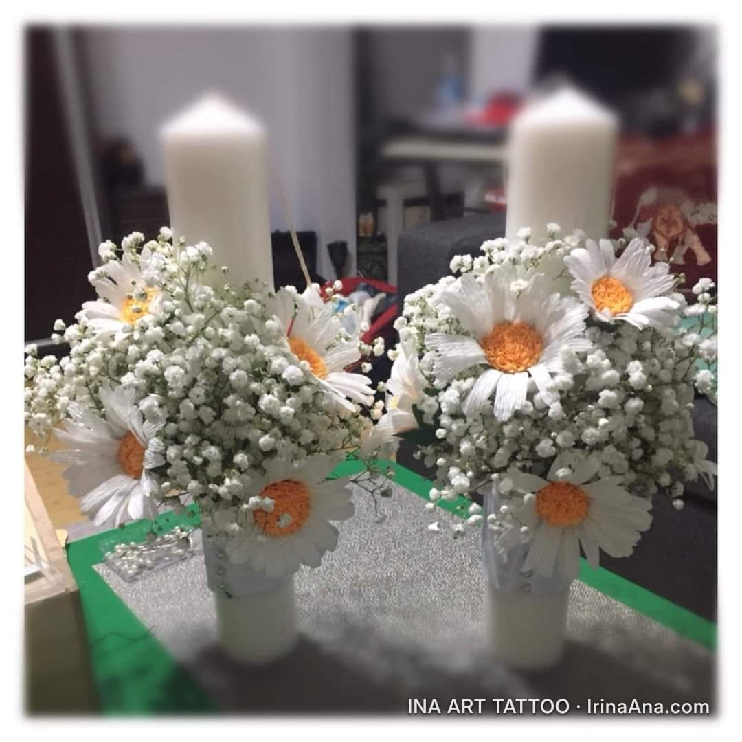 Two crepe paper floral arrangements, with white daisies and small white flowers, next to white candles. Blurred background.