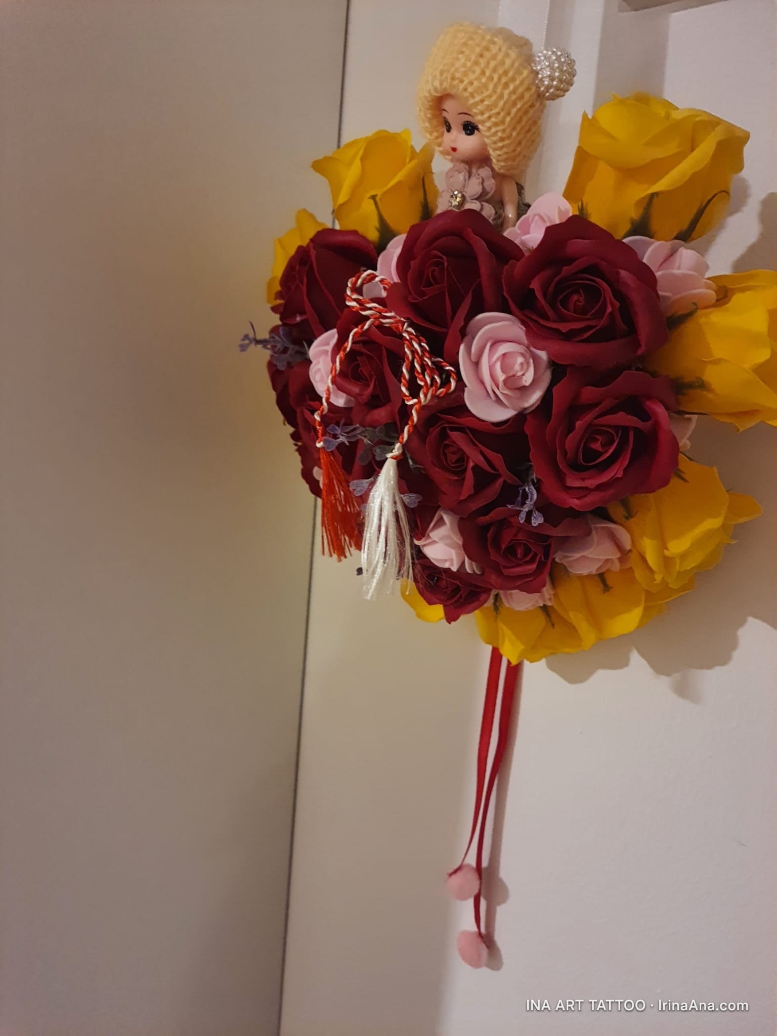 A doll with a yellow knitted hat, placed in a bouquet of red, pink, and yellow crepe paper roses.