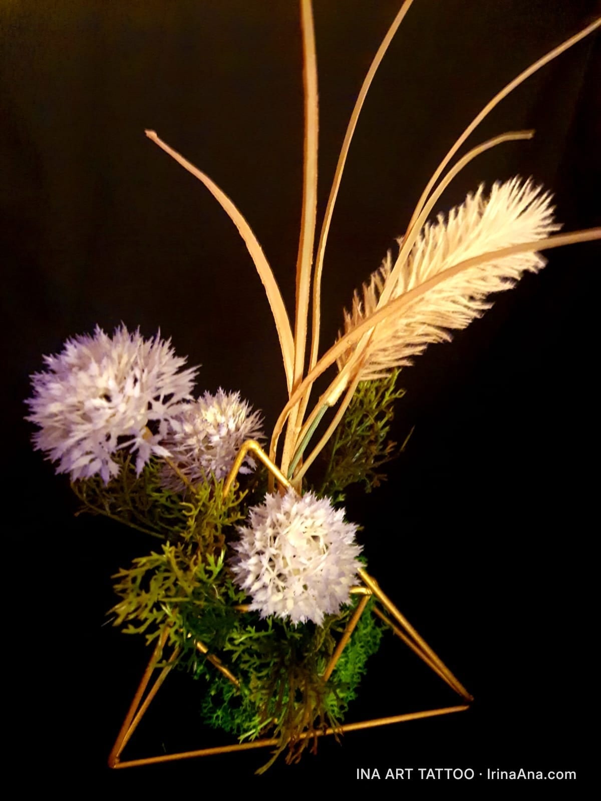 A handmade floral arrangement with white flowers, dried grass, and moss, set in a golden geometric structure, against a blac…