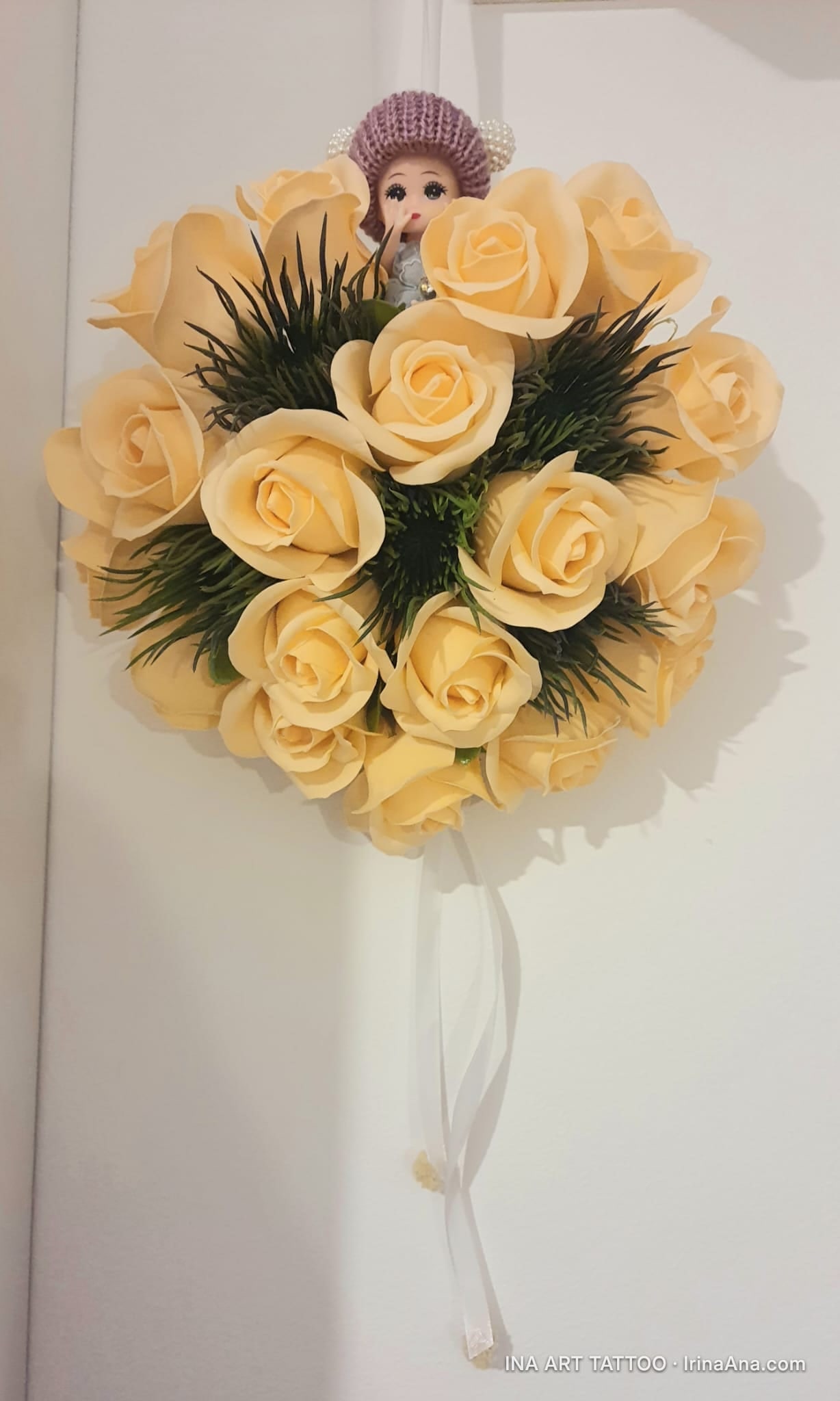 Round bouquet of yellow crepe paper roses, with a doll head wearing a purple hat in the center and a white ribbon hanging.