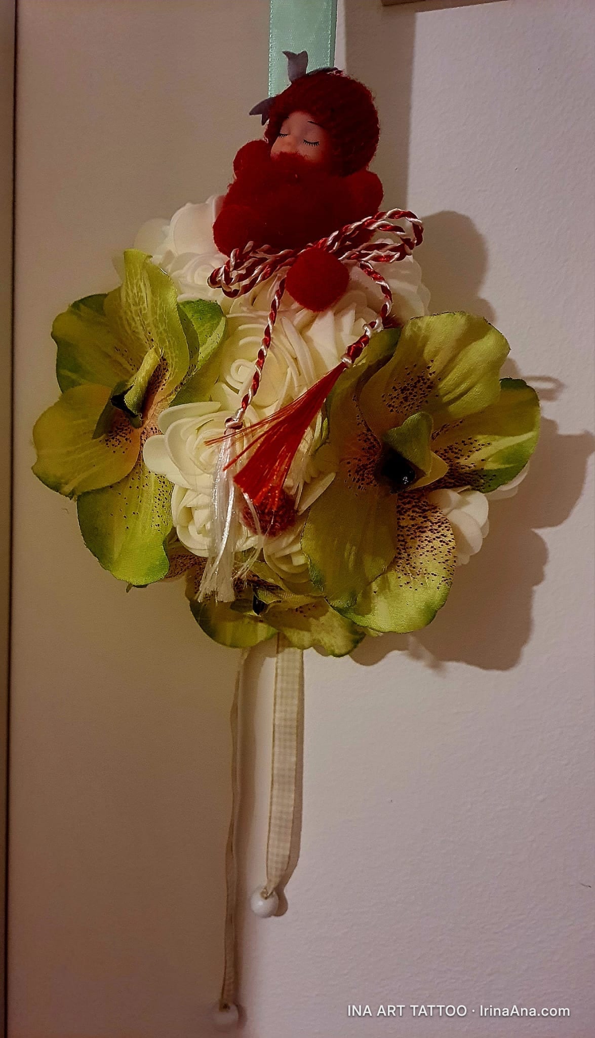 A floral mărțișor with green orchids and white flowers, topped with a red doll and red-and-white tassels.